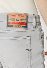 Jeans de color gris claro con una etiqueta de cuero que presenta texto en rojo y azul. La etiqueta dice "Diesel Industry" y destaca detalles sobre la calidad y el registro.