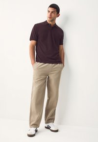 Tamsiai burgundiškos spalvos polo marškinėliai su subtiliu apykakliu, derinami su šviesiai smėlio spalvos kelnėmis. Balti sportbačiai su trimis juodomis juostelėmis ir pilku akcentu.