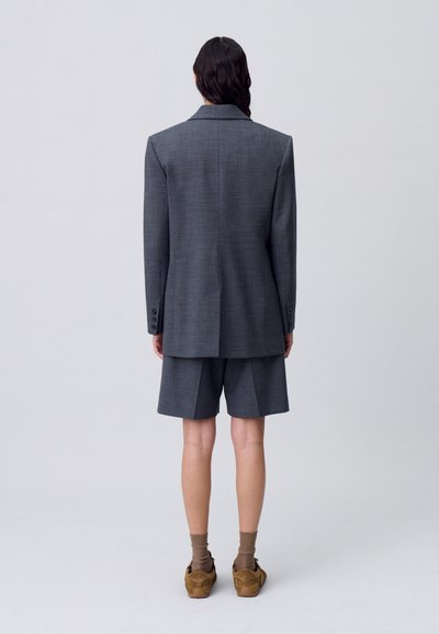 Blazer a scacchi grigi abbinato a pantaloni corti plissettati. Caratterizzato da spalline, polsini con tre bottoni e una vestibilità sartoriale. Indossato con calze marroni e scarpe.