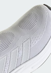 Grå atletisk sko med en texturerad stickad ovandel, som har släta överlägg och en vadderad vit sula. Subtila randmönster förhöjer designen.