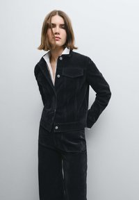 Giacca in velluto a coste nera con finiture in argento, caratterizzata da due tasche frontali e design con colletto, abbinata a pantaloni a gamba larga coordinati.