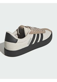 Bež platnena tenisica s crnim gumenim potplatom, kožnim naglaskom na peti i tri crne pruge sa strane. Zaobljeni vrh i teksturirani gornji dio.