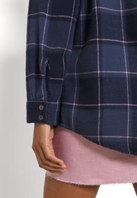 Camicia di flanella blu navy con motivo a quadri rosa, polsini con bottoni e orlo leggermente curvo, abbinata a una gonna di jeans rosa chiaro.