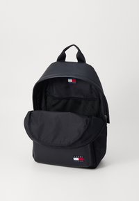 Sac à dos en nylon noir avec une forme arrondie, disposant d'une poche avant, d'un intérieur en mesh et d'un patch logo. Deux poignées pour le transport au niveau supérieur.