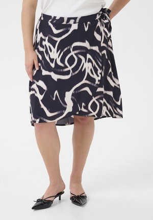 Mujer con una falda hasta la rodilla con un patrón abstracto en blanco y negro anudada en la cintura, combinada con zapatos de tacón bajo de punta negra con lazos.