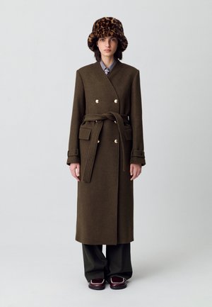 Cappotto di lana verde oliva con design avvolgente, bottoni dorati e cintura legata. Abbinato a un cappello a secchiello con stampa leopardata, pantaloni a gamba larga e scarpe marroni.