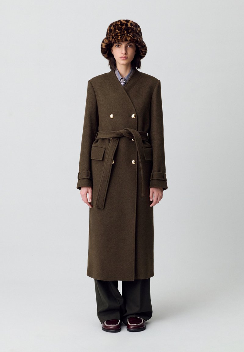 Cappotto di lana verde oliva con design avvolgente, bottoni dorati e cintura legata. Abbinato a un cappello a secchiello con stampa leopardata, pantaloni a gamba larga e scarpe marroni.