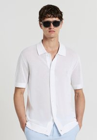 Weißes Kurzarmhemd aus Netzgewebe, mit einer Knopfleiste, klassischem Kragen und aufgerollten Ärmeln. Einheitlich hellblaue Hose.