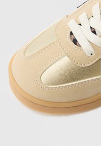 Chaussure de sport beige avec une tige texturée, des accents métalliques dorés, des lacets blancs et une semelle en caoutchouc couleur gomme. Dispose d'une bride auto-agrippante pour l'ajuster.
