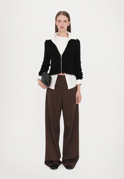 Zwarte geribbelde cropped sweater met rits over een witte blouse, gecombineerd met bruine wijde broek en een kleine zwarte clutch.