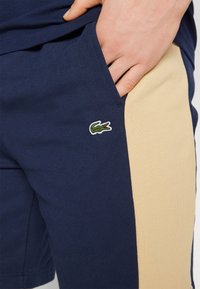 Shorts bleu marine avec un panneau latéral en tissu beige. Présente un logo de crocodile brodé en vert et rouge sur la poche gauche.