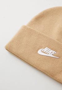 Bežs ribots adīts cepure ar saliektām malām, ar balti izšūtu Nike logo un svītru uz baltas fona.