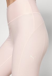 Jasnoróżowe legginsy z wysokim stanem, gładką teksturą i płaskimi szwami. Zawierają małe białe logo Pumy na lewej udzie.