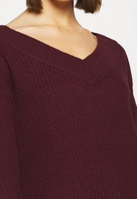 Maglione bordeaux lavorato a maglia con scollo a V, caratterizzato da un tessuto testurizzato e una vestibilità ampia. È visibile un particolare motivo a maglia grossa.