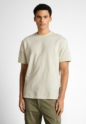 Young man standing facing forward wearing light beige short-sleeve T-shirt and olive green cargo pants against plain white background.