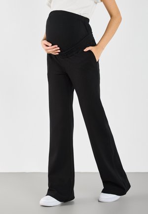 Personne enceinte portant un pantalon de maternité noir et des baskets blanches, tenant le ventre d'une main, l'autre main dans la poche sur un fond uni.