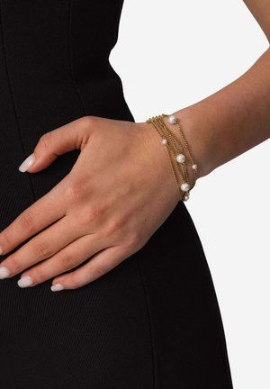 Goldkette Armband mit mehreren Strängen und weißen Perlen, getragen an einer Hand vor einem schwarzen strukturierten Hintergrund.