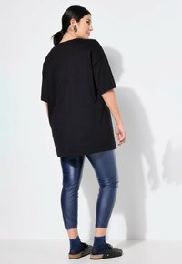 Zwarte oversized t-shirt met korte mouwen, gecombineerd met glanzende blauwe leggings. Zwarte schoenen en blauwe glinsterende sokken. Model staat met de rug naar de camera.