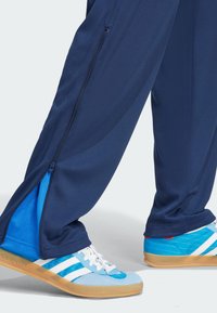Navy athletic pants with mesh texture, zippered side vents, paired with blue and white sneakers featuring horizontal stripes and gum sole.