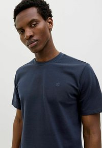 Tengerészkék crew neck póló rövid ujjakkal, puha anyagból készült, diszkrét logóval a bal felső sarokban. Sima textúra.