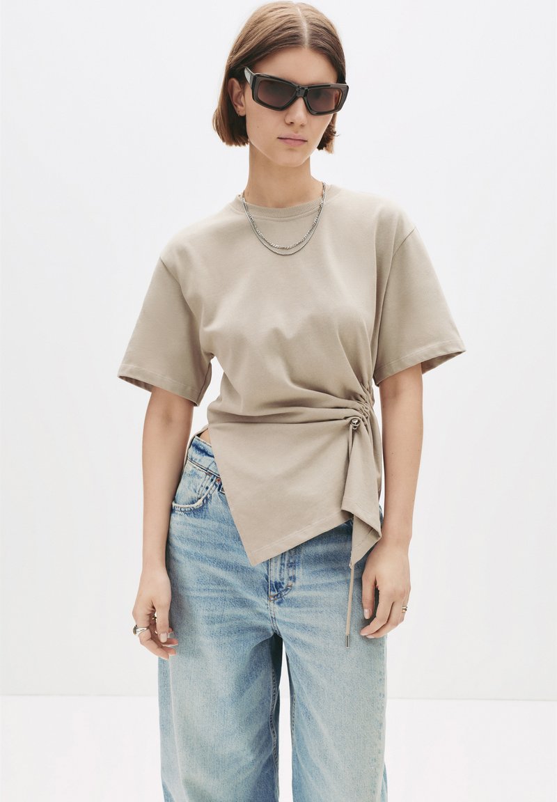 Kvinde iført beige kortærmet top med sidesløjfe, lyseblå jeans, rektangulære solbriller og lagdelte sølvkæder, stående mod hvid baggrund.