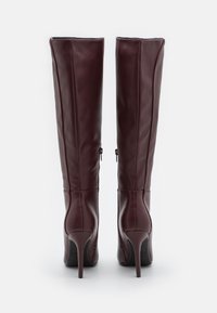 Bottes en cuir bordeaux montantes avec une texture lisse, des talons aiguilles hauts et des fermetures éclair sur les côtés pour un enfilage facile.