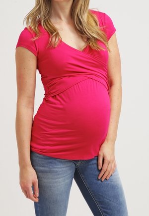 Femme enceinte portant un haut cache-cœur rose vif et un jean bleu, debout devant un fond clair uni.