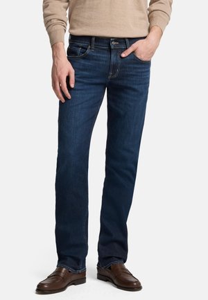 Uomo con jeans blu scuro a gamba dritta, maglione beige e mocassini di pelle marrone, una mano in tasca.