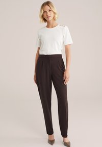 O femeie stă cu încredere, purtând un top alb cu mânecă scurtă și pantaloni eleganți de culoare maro închis, cu tocuri elegante și machiaj discret.