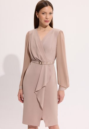 Kobieta w beżowej sukience kopertowej z dekoltem w kształcie litery V, w talii z paskiem, z długimi przezroczystymi bufkami na rękawach i falbanką z przodu.