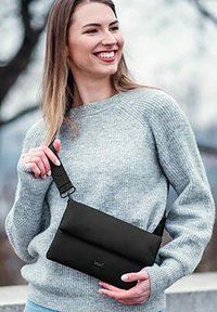 Černá látková crossbody taška s překlopným designem a nastavitelným popruhem, kterou drží osoba v svetru s jemnou šedou texturou.