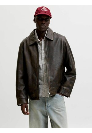 Osoba o ciemnej karnacji ubrana w czerwoną czapkę, brązową skórzaną kurtkę, koszulę w kratę zapinaną na guziki oraz jasnoniebieskie dżinsy na jednolitym tle.