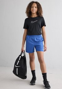 Noor inimene seisab mustas Nike särk, sinistes Nike lühikestes pükstesse, mustades Nike sokkides ja kingades, hoides musta Nike tõmbluku kotti.