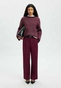 Femme avec de longs cheveux bouclés portant un pull texturé bordeaux et un pantalon assorti, tenant un sac à main noir, se tenant devant un fond blanc.