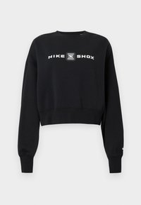 Černá zkrácená mikina vyrobená z měkké látky, s dlouhými rukávy a kulatým výstřihem. Uprostřed je na přední straně bílé logo Nike Shox.