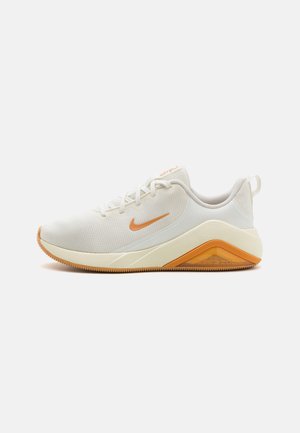 Chaussure de course Nike blanche et beige avec un swoosh et des accents de semelle orange, dessus en mesh, fermeture à lacets et soutien talon amorti Air Zoom.