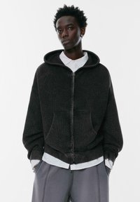 Czarny, żeberkowany hoodie na zamek z teksturowanej tkaniny, z dużym kapturem, przednimi kieszeniami i luźnym krojem, nałożony na jasną koszulę z kołnierzykiem.