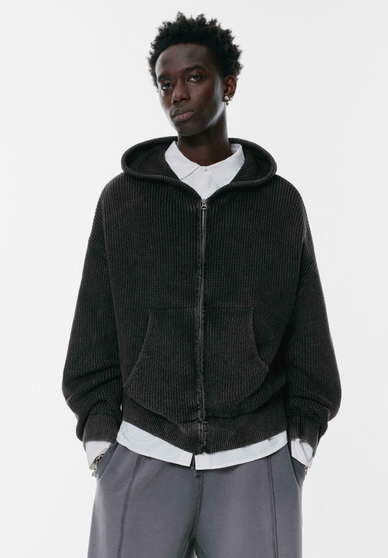 Czarny, żeberkowany hoodie na zamek z teksturowanej tkaniny, z dużym kapturem, przednimi kieszeniami i luźnym krojem, nałożony na jasną koszulę z kołnierzykiem.