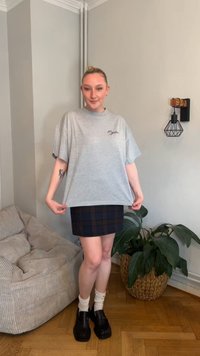 Grå oversized t-skjorte med korte ermer, med en liten logo. Kombinert med et rutete miniskjørt i mørke toner og svarte loafers.