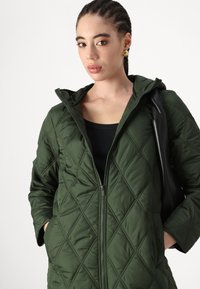 Grüne Steppjacke mit Kapuze, ausgestattet mit einem Frontreißverschluss, zwei Seitentaschen und einem strukturierten Rautenmuster. Schwarzes Oberteil darunter, kombiniert mit einer Tasche.