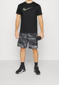 Camiseta negra de Nike con logo de camuflaje, combinada con pantalones cortos de camuflaje gris que presentan un logo blanco de Nike. Zapatillas deportivas negras y botella de agua.