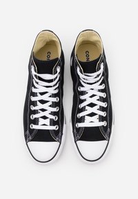Paire de baskets montantes noires en toile avec lacets blancs, bouts en caoutchouc blanc et coutures visibles sur fond clair.