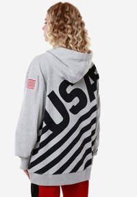 Felpa grigia con grande scritta nera "USA" e strisce ondulate sul retro, con un piccolo patch della bandiera americana sulla manica sinistra.