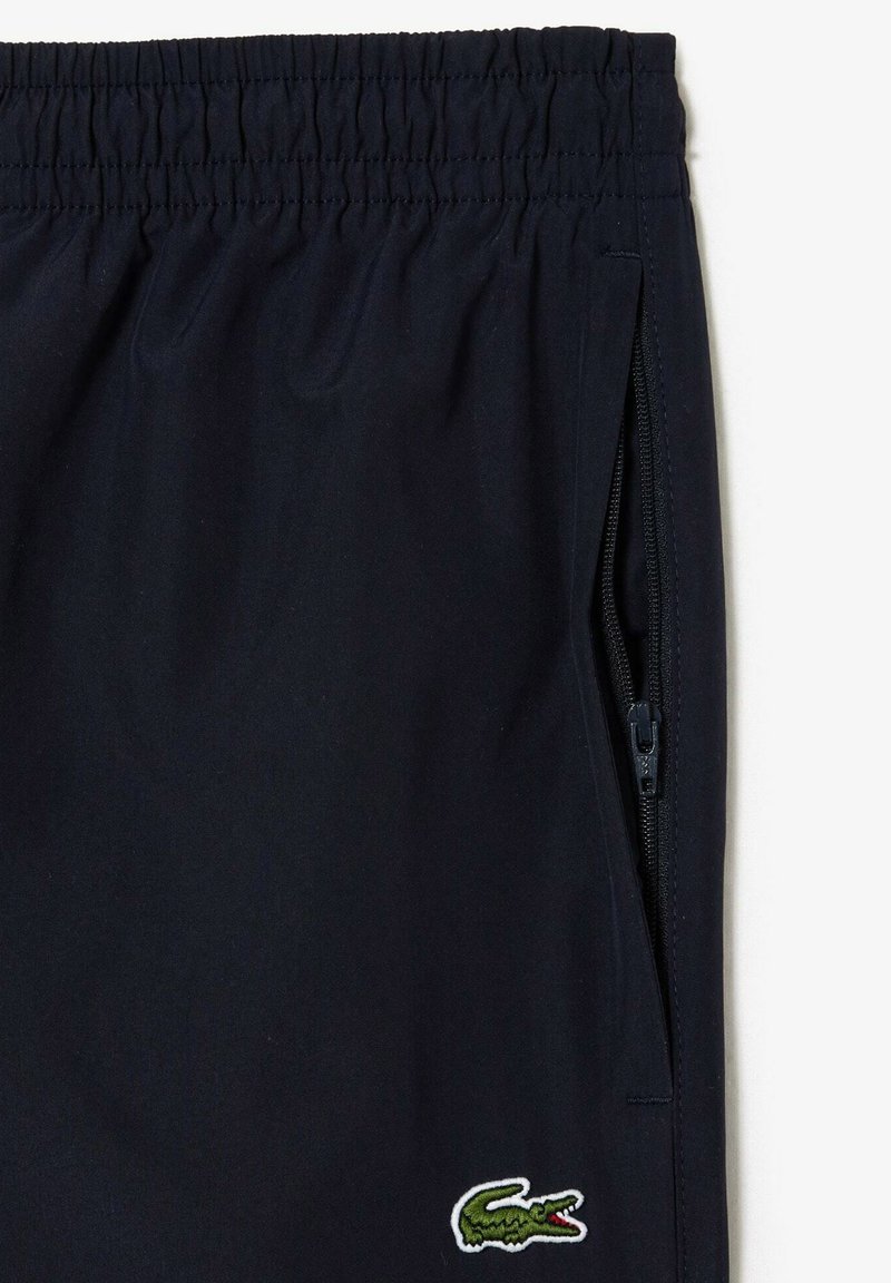 Shorts bleu marine avec taille élastique, poche zippée sur le côté et logo crocodile vert Lacoste près de l'ourlet.