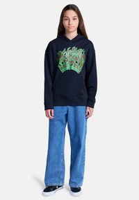 Fiatal személy áll, fekete kapucnis pulóvert visel zöld lángmintával és kék széles szárú farmert, egyszínű fehér háttér előtt.