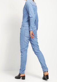 Ljusblå denimsoverall med lång ärm, figurnära passform, två framsid fickor och avsmalnande ben, parat med svarta klackskor.