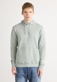 Jongeman in een lichtgrijze hoodie met een voorzak en een blauwe spijkerbroek, staand tegen een effen witte achtergrond.