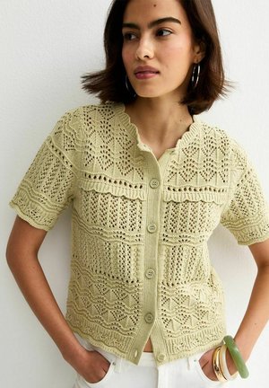 Giovane donna che indossa un top beige a maniche corte con bottoni e motivi in pizzo intricati, pantaloni bianchi e diversi braccialetti al braccio.