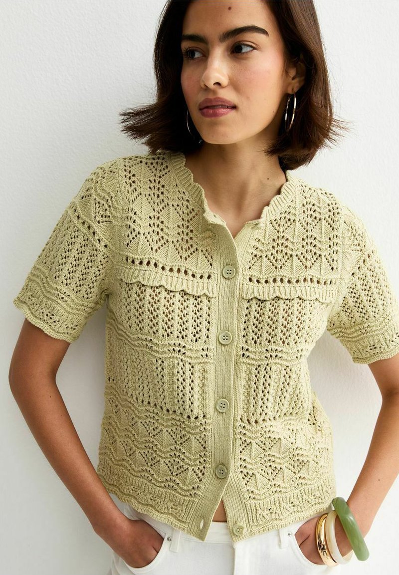 Jeune femme portant un haut en maille beige à manches courtes boutonné avec des motifs de dentelle complexes, un pantalon blanc et plusieurs bracelets à son bras.