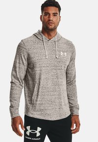Sudadera gris, texturizada, de manga larga con un bolsillo frontal y cordones blancos. Logo de Under Armour en el pecho, combinada con joggers oscuros.
